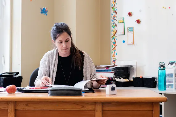   Substitute professional sitting at a desk, reading a book in a classroom with colorful decorations